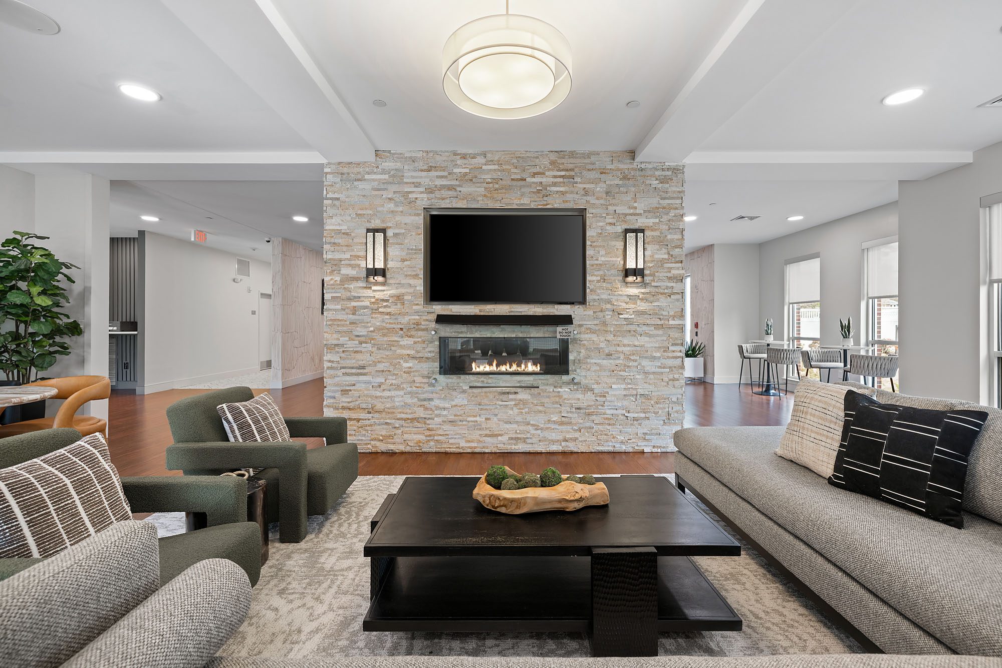 Modern Ken-Caryl Ranch living room with stone accent wall, mounted TV, and fireplace. Neutral-toned furniture includes a gray sofa, two green armchairs, and a black coffee table with wood centerpiece. Dining area visible in the background.