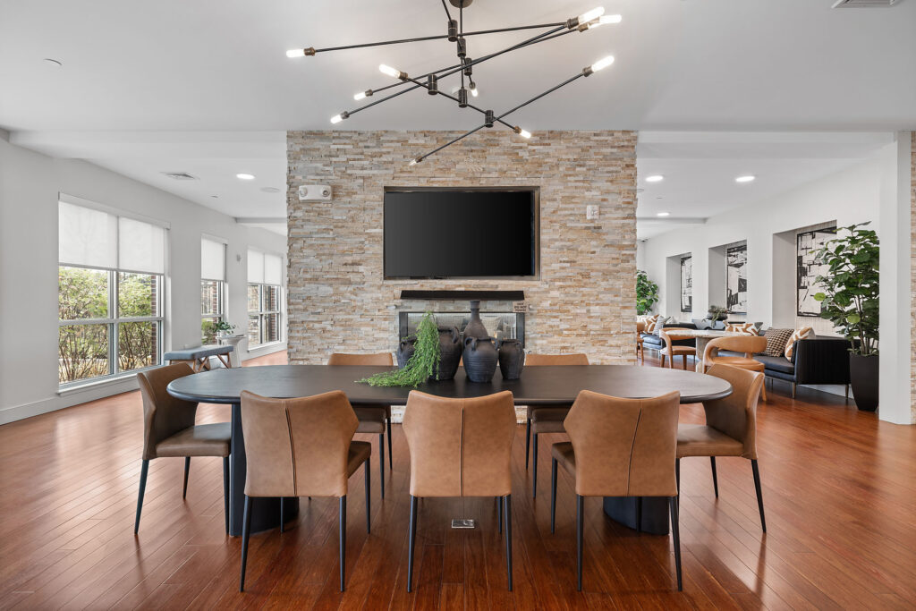Modern dining area with a round table, tan chairs, decorative vases, and a large stone accent wall featuring a mounted TV and fireplace; open layout with wood floors and large windows.