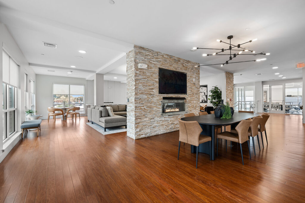 Modern open-concept lounge with wood floors, a stone accent wall with a fireplace and mounted TV, a dining table with chairs, and large windows providing natural light. Sofa and seating area are visible in the background.