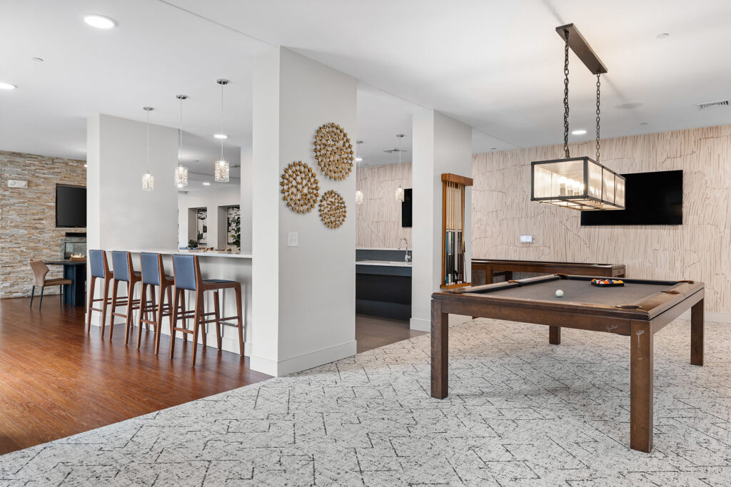Modern recreation room with a pool table, flat-screen TV, decorative wall art, and a bar area with high chairs; features wood floors, pendant lighting, and contemporary décor.