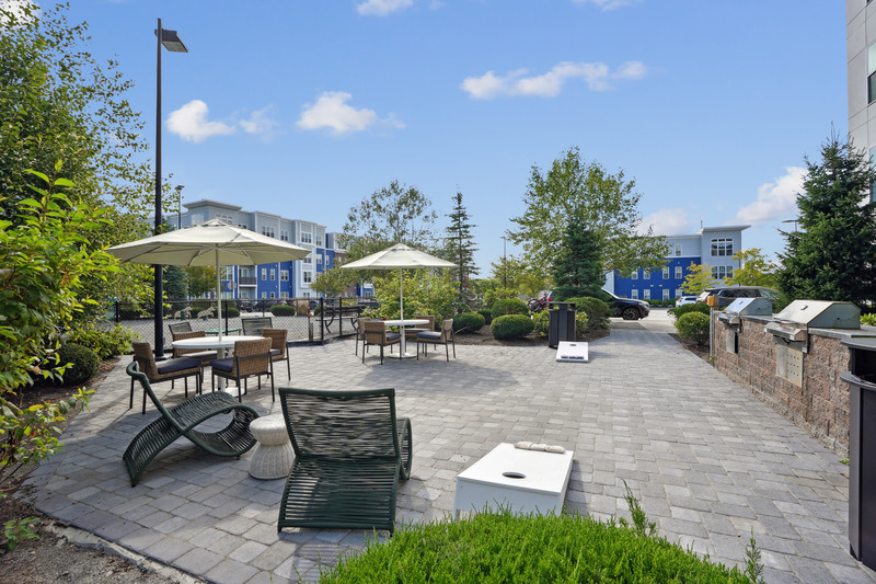 Outdoor patio in Littleton, CO with lounge chairs, tables with umbrellas, and cornhole boards on a stone-paved area, surrounded by shrubs and trees. Modern apartment buildings rise in the background under a blue sky.