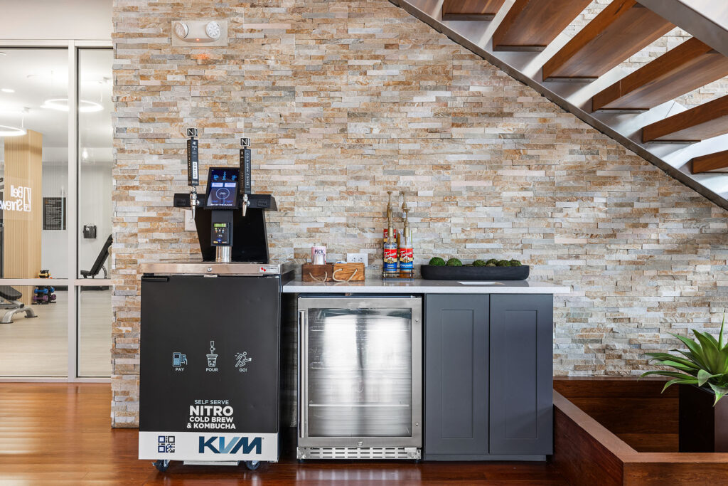 A modern beverage station under a staircase features a nitro cold brew and kombucha dispenser, a mini fridge, gray cabinets, glass jars with straws, and a bowl of limes, all against a stone accent wall.