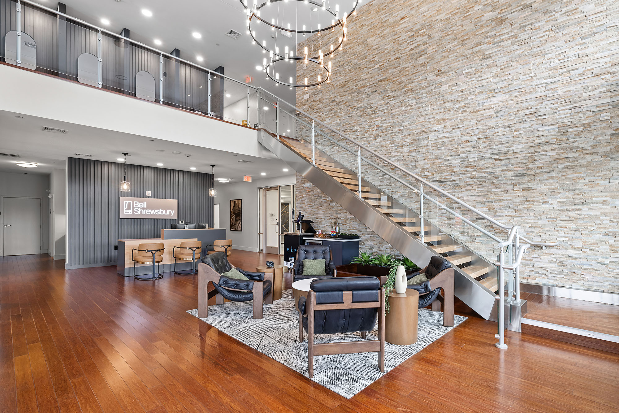 A modern office lobby in Littleton CO with wood floors, a stone accent wall, a large staircase, seating area with chairs and tables, and a reception desk featuring a Bell Shrewsbury sign in the background.