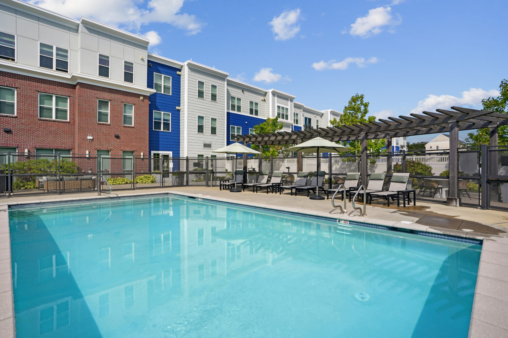 Outdoor swimming pool with lounge chairs and umbrellas beside modern apartment buildings in red, white, and blue in Littleton CO. A metal pergola provides shade, and the sky is blue with scattered clouds.