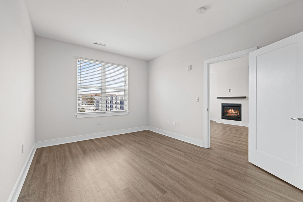 A bright, empty room with light wood flooring, white walls, a window with blinds, and an open door leading to another room with a lit fireplace.