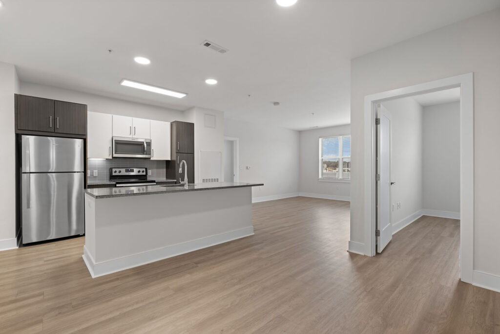 Modern apartment kitchen with stainless steel appliances, white cabinets, and a breakfast bar, opening to a spacious living area with wood floors and a doorway leading to a bright bedroom.