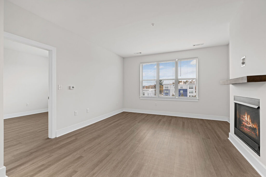 A bright, empty living room with light wood flooring, white walls, a large window with blinds, a modern built-in fireplace, and a doorway leading to another room.