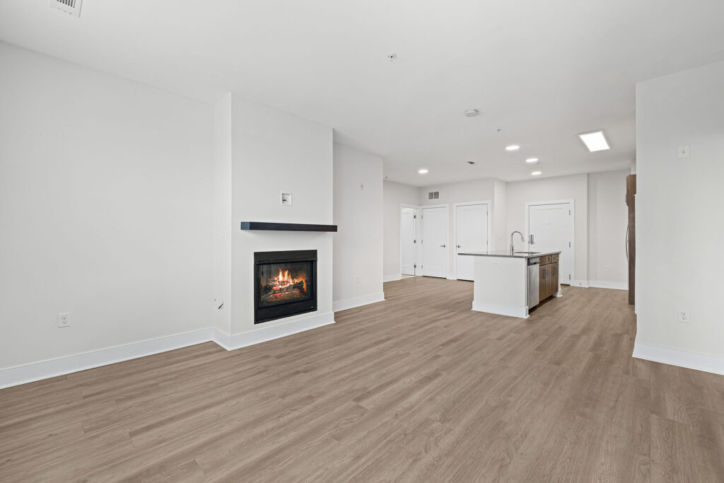 Modern, unfurnished open-concept living space with light wood floors, a corner fireplace, white walls, and a kitchen area featuring an island and stainless steel sink in the background.