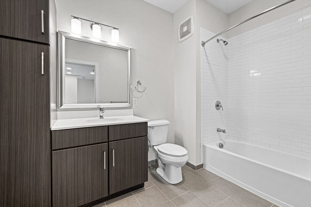 Modern bathroom with dark wood vanity, white countertop, large mirror, toilet, and a white bathtub with shower and curved curtain rod. Walls are light-colored, floor is tiled, and lighting is above the mirror.