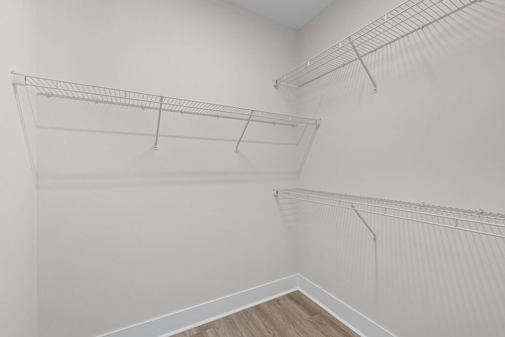 Empty walk-in closet with white wire shelves on three walls, light-colored walls, and wood-style flooring. The space is clean and well-lit.