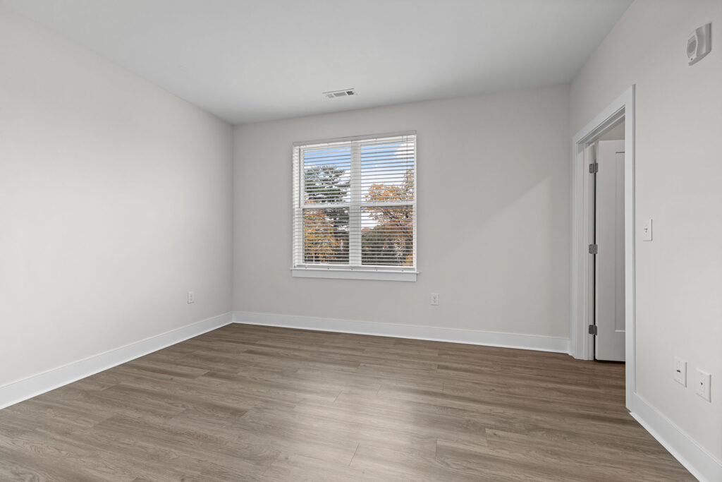 A bright, empty room with light-colored walls, a large window with white blinds, and wood-look flooring. Sunlight streams through the window, and there is an open door on the right side.