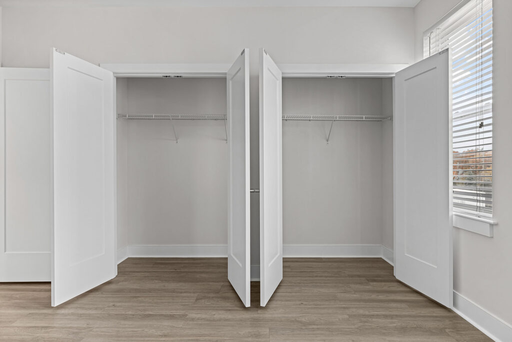 Two open white closets with empty wire shelves in a bright room with light gray walls, white trim, and wood-look flooring. Sunlight streams in through a window with white blinds on the right.