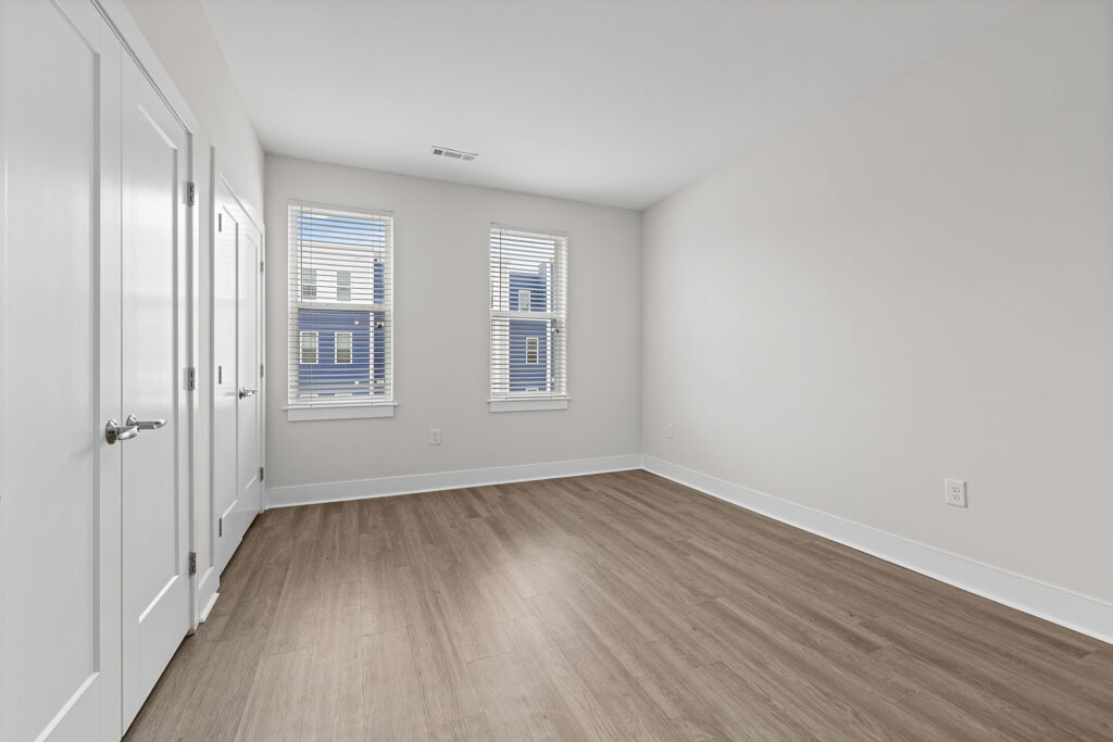 A bright, empty room with light-colored walls, two windows with white blinds, double closet doors, and wood-style flooring. Natural light fills the space, giving it a clean and modern appearance.