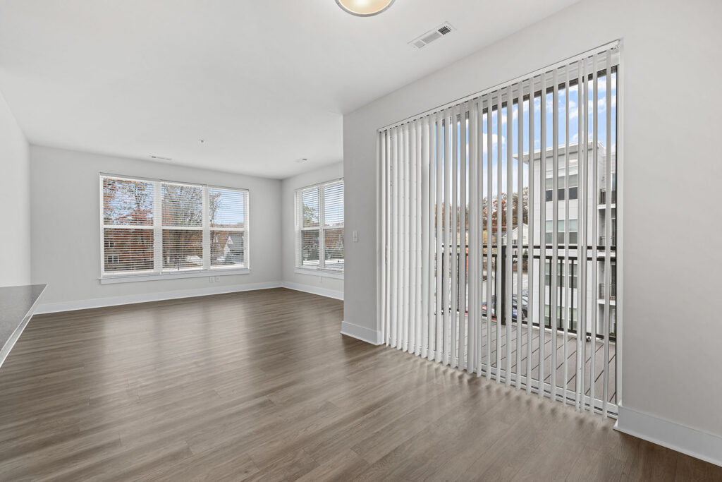 Bright, empty room with large windows, white walls, wood-style flooring, and vertical blinds covering a sliding glass door that opens to a balcony with a view of trees and nearby buildings.