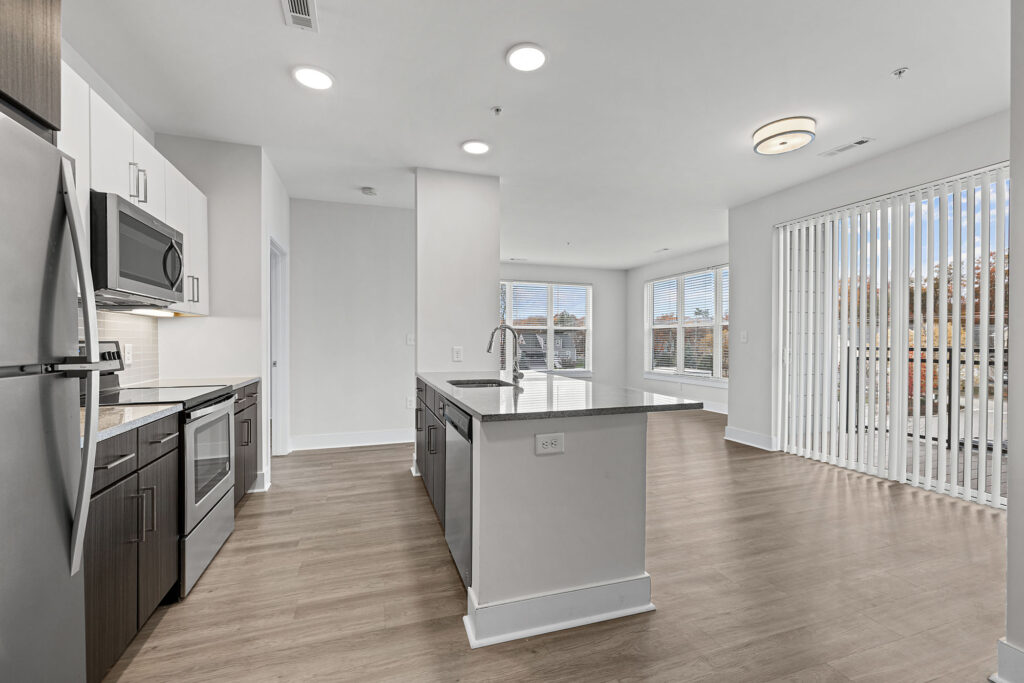 Modern kitchen with stainless steel appliances, an island with a sink, white cabinets, light wood flooring, and large windows with vertical blinds letting in natural light into an open living space.