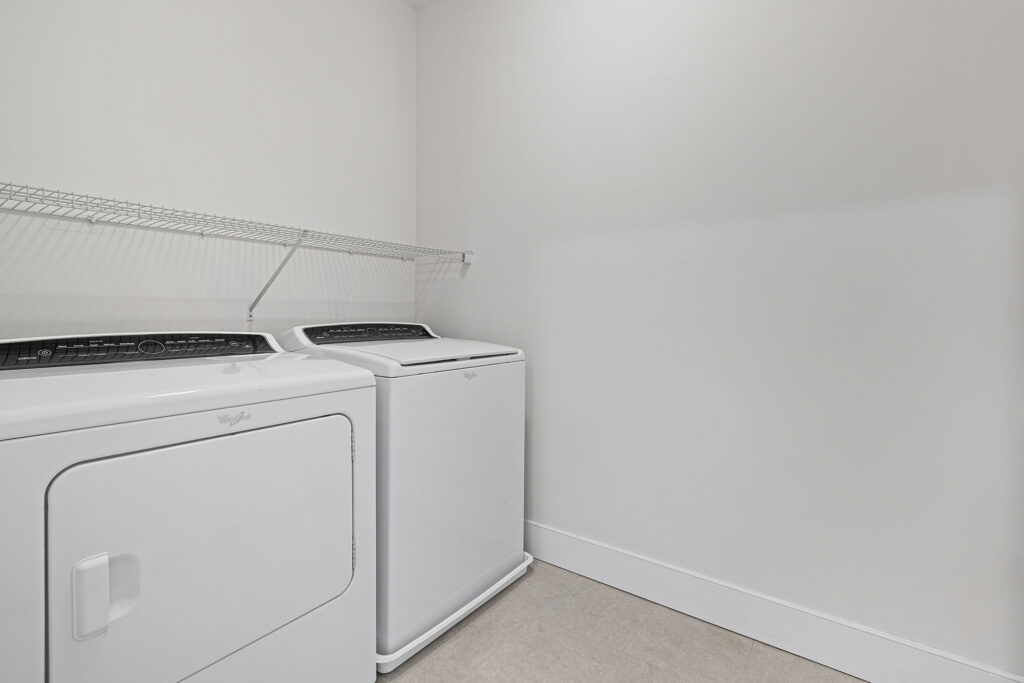 A laundry room with a white washer and dryer set side by side beneath a white wire shelf, against plain light-colored walls and floor.