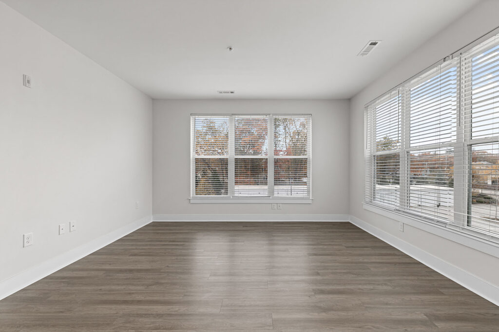 Bright, empty room with light gray walls, large windows with white blinds, and wood-style flooring. Natural light fills the space, and trees with autumn colors are visible outside.