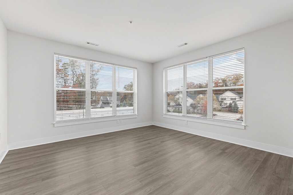 Bright, empty room with large corner windows, white walls, and light wood flooring. Trees and houses are visible outside through the blinds, allowing natural daylight to fill the space.
